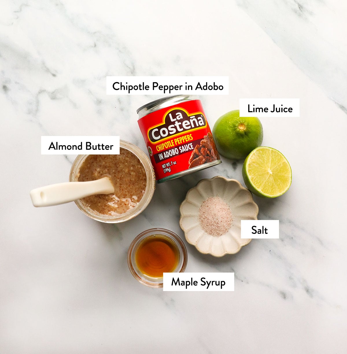 jar of almond butter, chipotle peppers, salt, lime, and maple syrup on a white surface.
