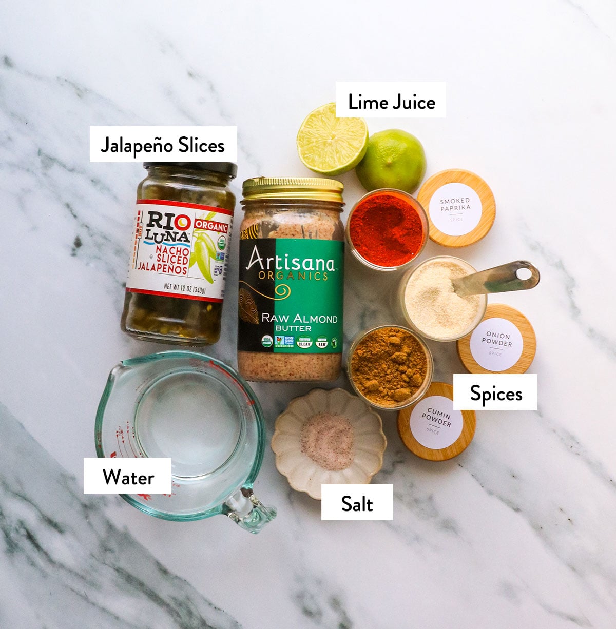 jar of almond butter, jalapeno slices, pantry spices, and a lime on a white surface.