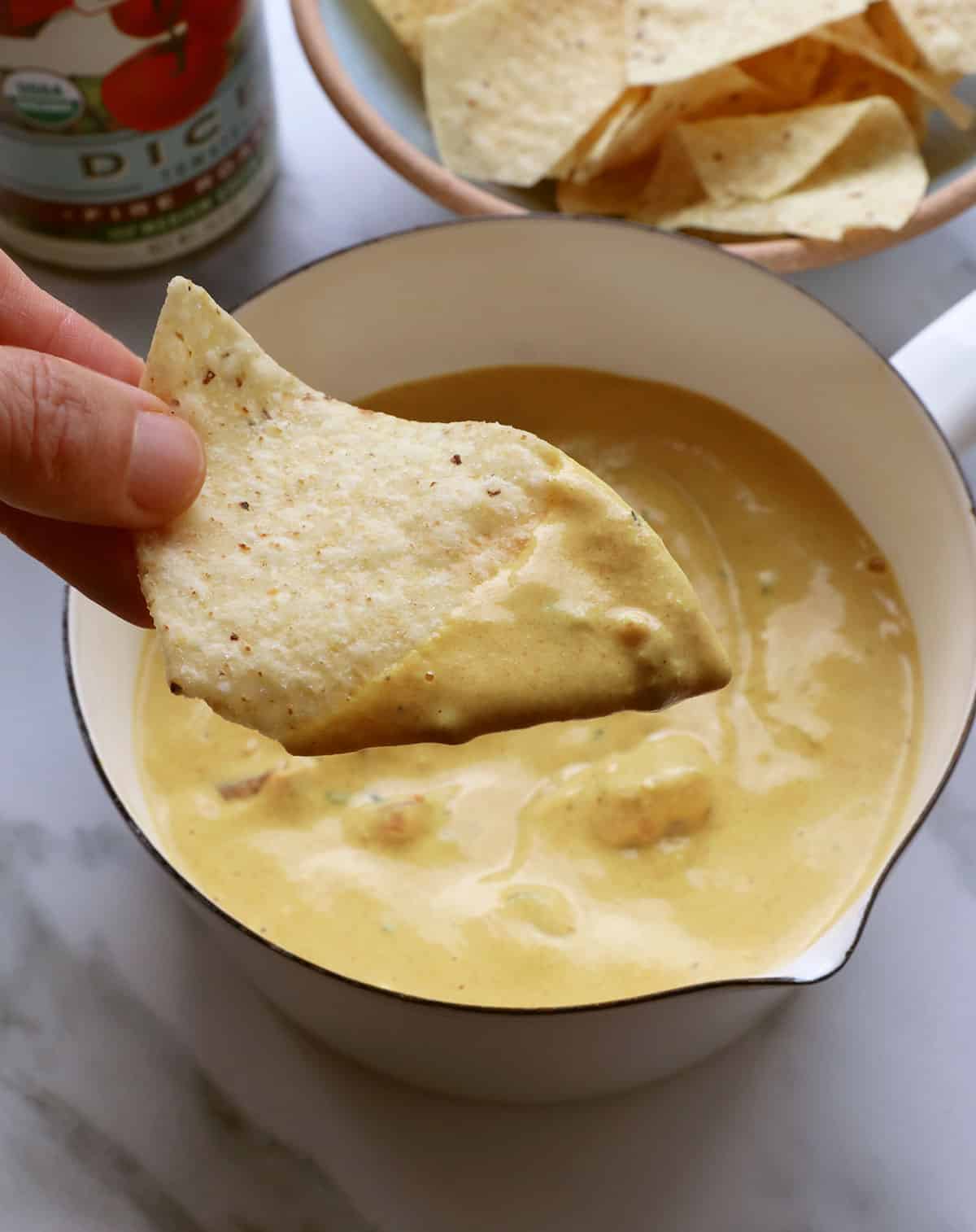 fingers holding a chip with dairy-free queso on it over a pan.