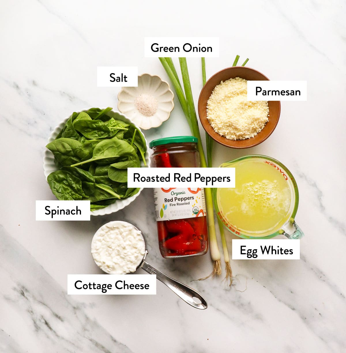 Baby spinach, jarred roasted red peppers, egg whites, cottage cheese, and Parmesan in bowls on a white surface. 